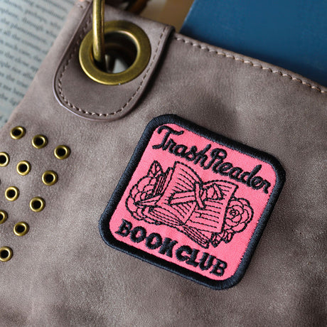 Pink machine embroidery patch with 'Trash Reader Book Club' text on a brown leather bag.