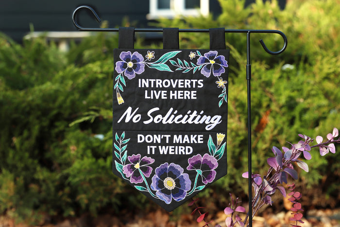 Decorative garden flag with machine embroidery floral design and text about introverts and soliciting, attached to a stand outdoors.
