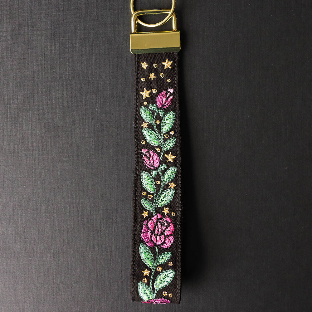 Digitally downloadable machine embroidery design of a floral wristlet strap with pink roses, green leaves, and small gold stars on black fabric.