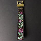 Digitally downloadable machine embroidery design of a floral wristlet strap with pink roses, green leaves, and small gold stars on black fabric.