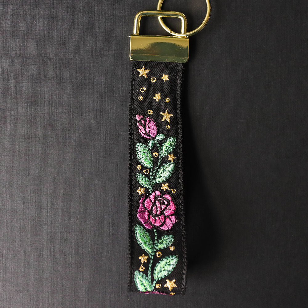 Digitally downloadable machine embroidery design of pink flowers with green leaves and gold stars on a black wristlet strap with gold hardware.