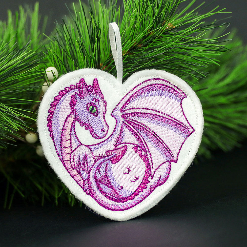 Heart-shaped white fabric ornament with embroidered curled purple dragon with green eyes and detailed wings hanging from a pine branch.
