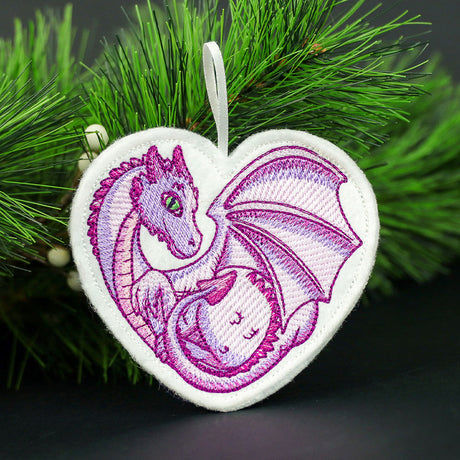 Heart-shaped white fabric ornament with embroidered curled purple dragon with green eyes and detailed wings hanging from a pine branch.