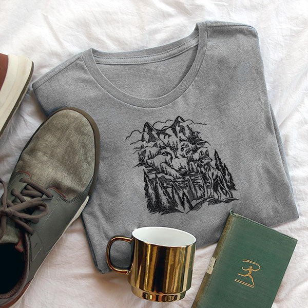 Gray sweatshirt with machine embroidered wolf and mountain design next to a book, mug and shoes.