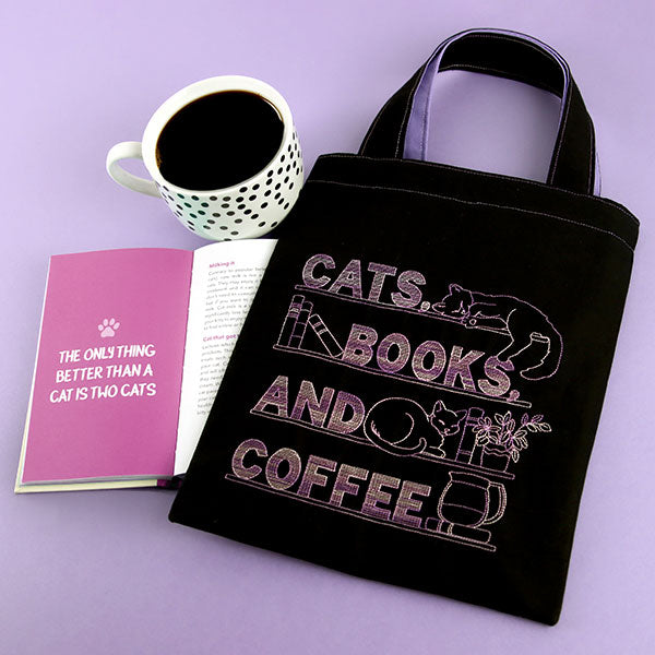 Black tote bag with machine embroidered 'Cats, Books, and Coffee' design, next to a mug and book on a purple background