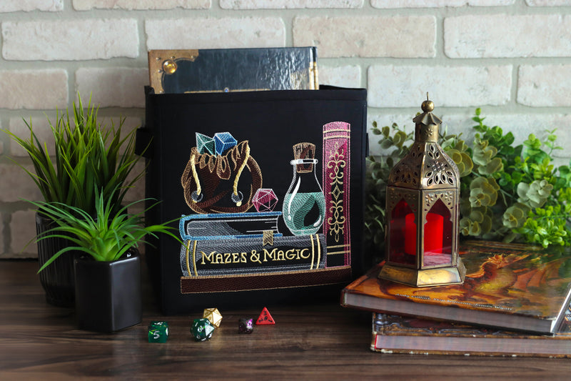 Black tote bag with machine embroidery 'Mazes & Magic' bookshelf design on a wooden surface with decorative items.