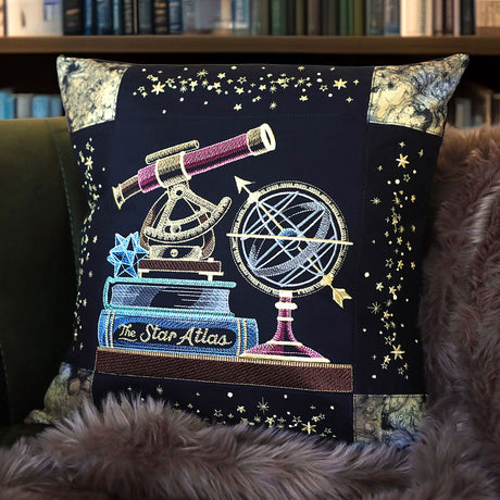Black pillow embroidered with celestial  bookshelf panel machine embroidery design featuring a telescope, star atlas, and globe on a dark background.
