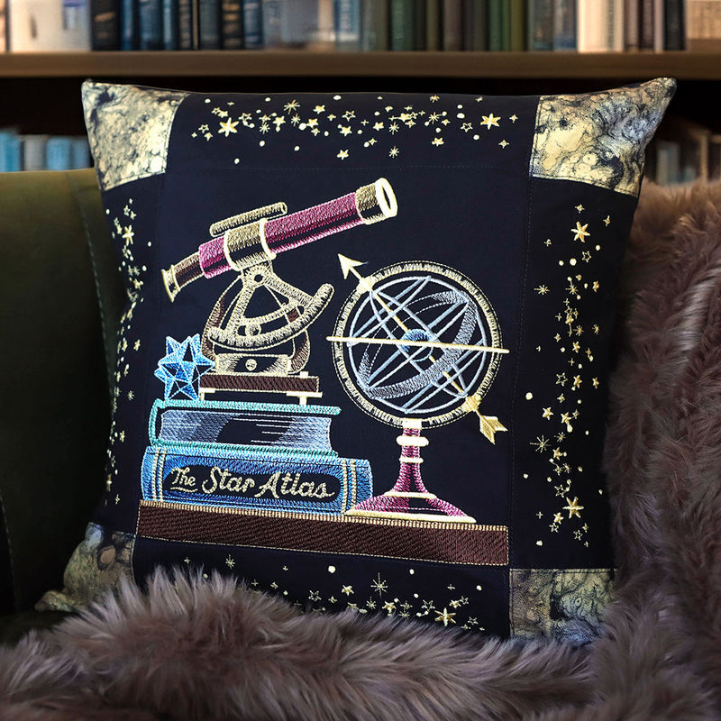 Black pillow embroidered with celestial  bookshelf panel machine embroidery design featuring a telescope, star atlas, and globe on a dark background.