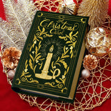 A green book titled "A Christmas Carol" with gold embroidery of a lit candle and holly leaves is placed on a red surface surrounded by gold and copper holiday decorations.