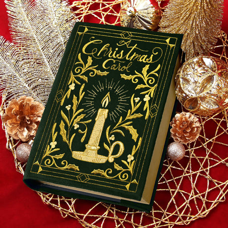 A green book titled "A Christmas Carol" with gold embroidery of a lit candle and holly leaves is placed on a red surface surrounded by gold and copper holiday decorations.