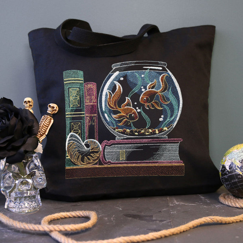 Black tote bag with colorful embroidery of two goldfish in a bowl, two stacked books, and a seashell, placed on a dark surface with a skull vase and globe nearby.