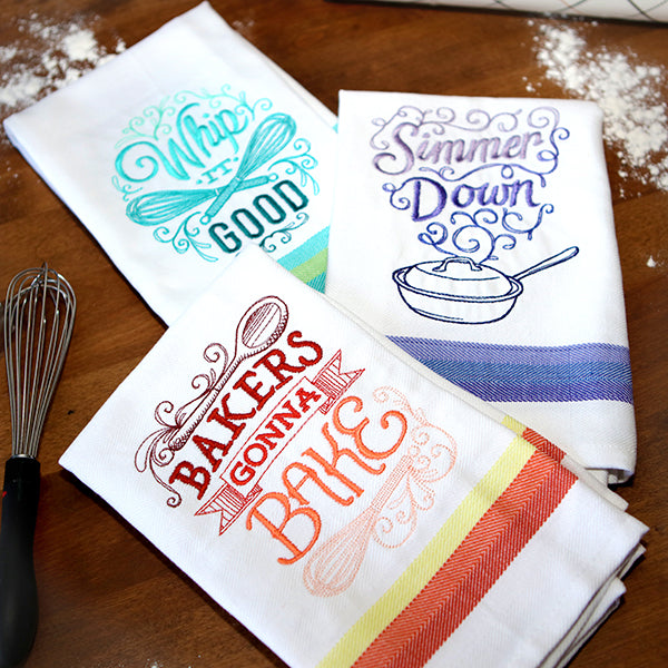 Three kitchen towels with colorful sassy  machine embroidered kitchen sayings on a wooden counter top.