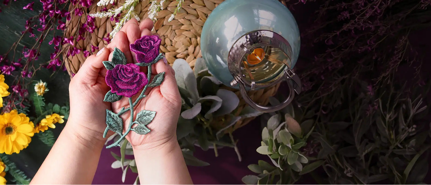 Hand holding a decorative machine embroidery flower with a lantern and flowers in the background