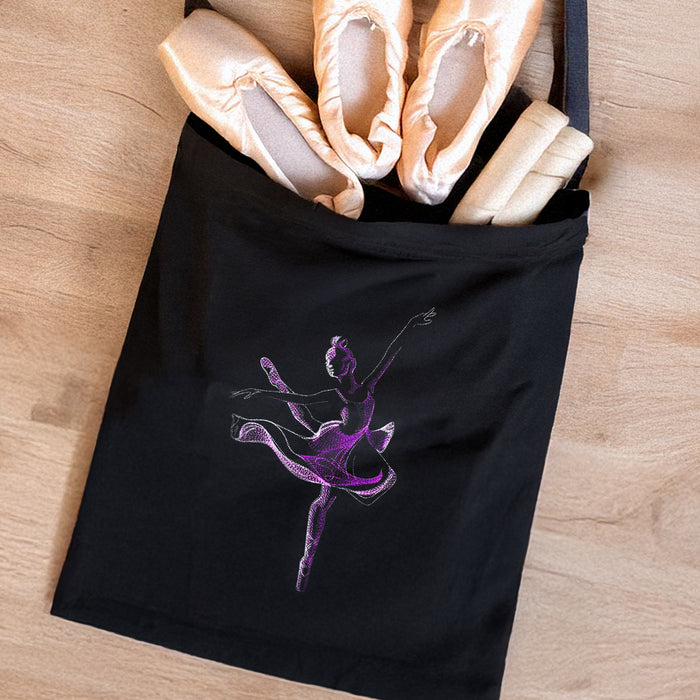 Black tote bag with a purple machine embroidered ballet dancer design, containing ballet shoes on a wooden surface.