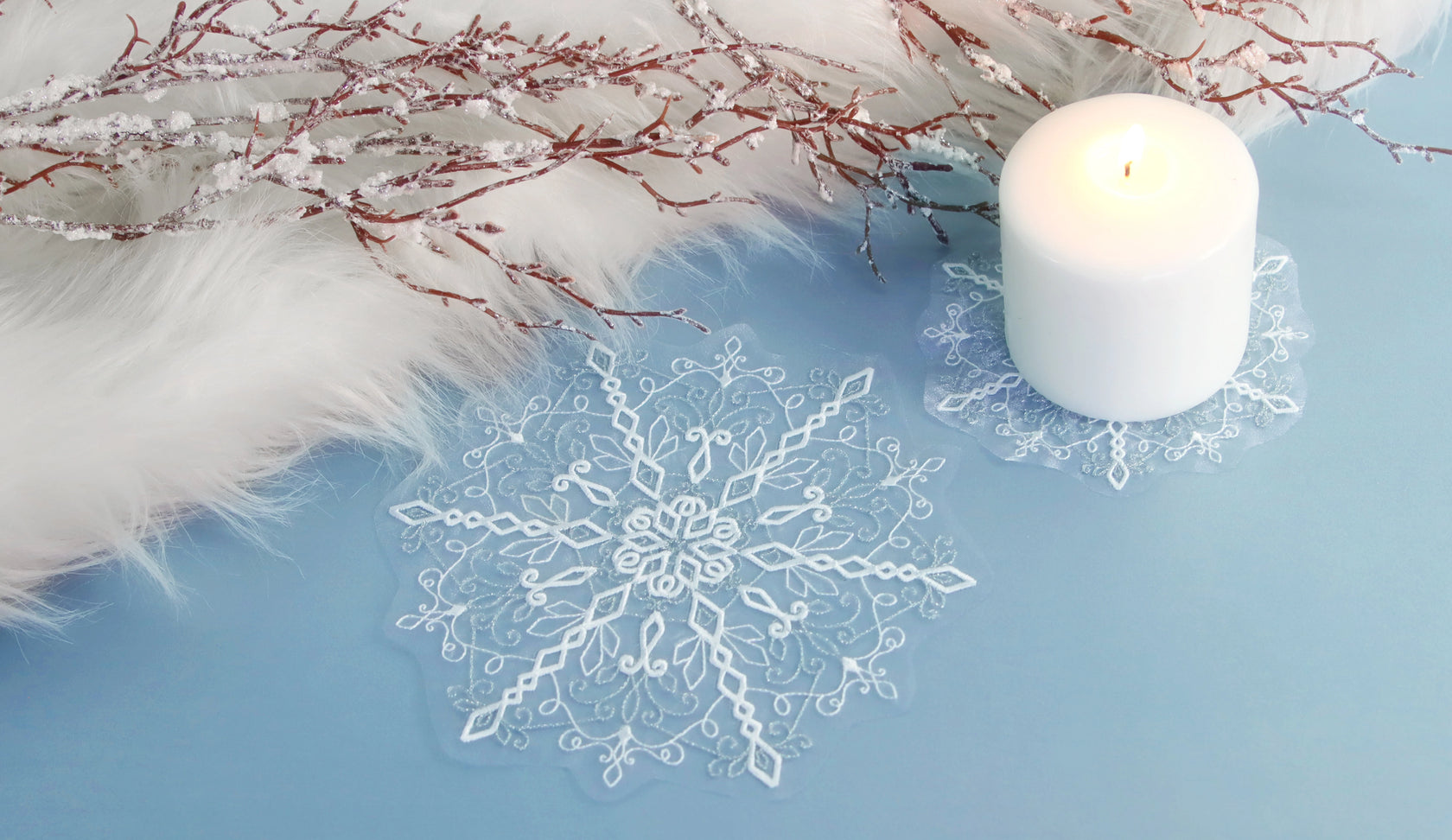Machine embroidered freestanding lace snowflake on light blue surface with a white candle and frosted branches