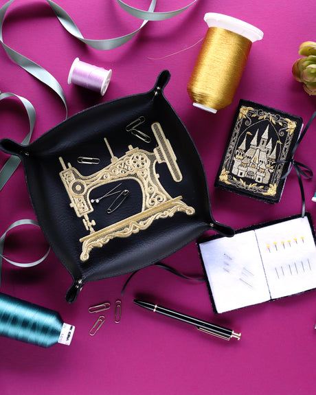 Machine embroidery sewing machine on a black catch all tray surrounded by sewing supplies and a machine embroidery castle needlebook.