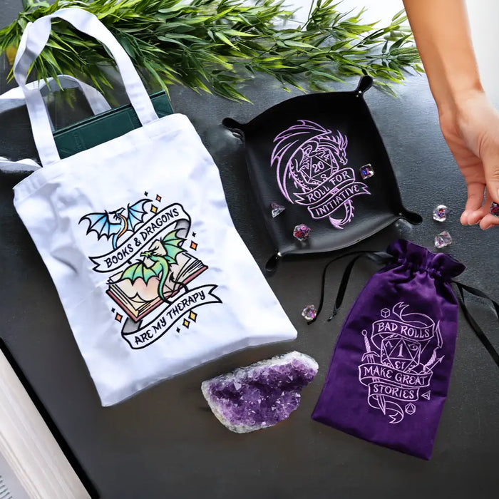 Three fantasy-inspired machine embroidery projects including a white tote bag with dragon book design, a black dice tray with dragon embroidery, and a purple dice bag with gaming-themed design.
