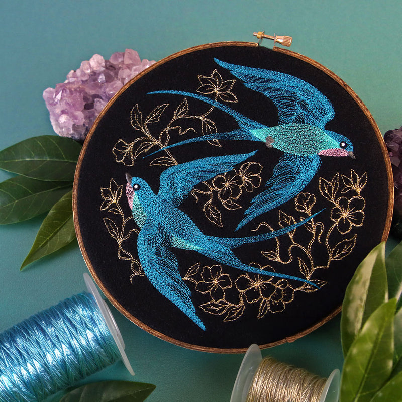 Embroidery hoop featuring two blue swallows stitched on black fabric with gold floral details, surrounded by thread spools, greenery, and purple crystals.