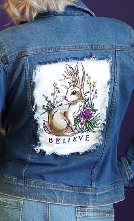 Denim jacket with a machine embroidery jackalope and flowers and the word 'BELIEVE'.
