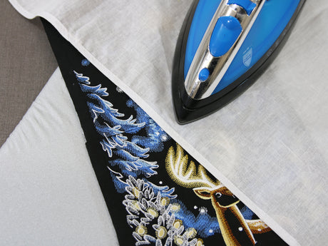 Ironing a fabric with a machine embroidered winter deer scene design on a white surface.