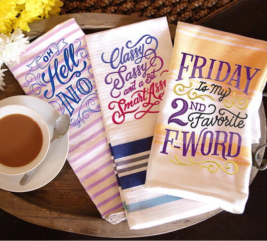 Set of decorative kitchen towels with humorous sayings on a wooden tray with a cup of coffee.