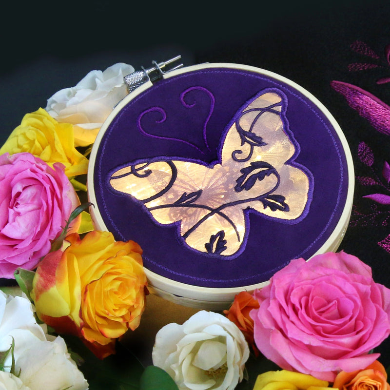 An machine embroidered organza butterfly on purple fabric in a hand embroidery hoop surrounded by flowers.