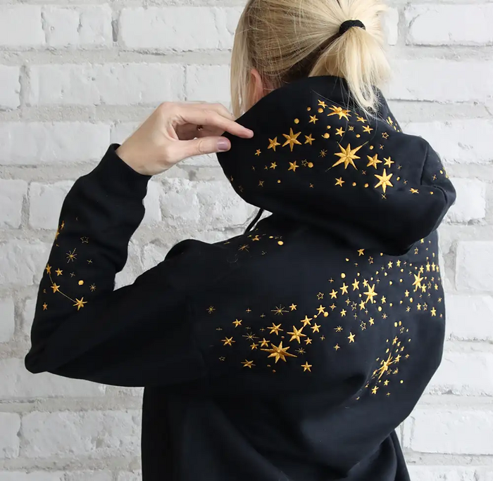 Person wearing a black hoodie with machine embroidery gold star designs against a white brick wall.
