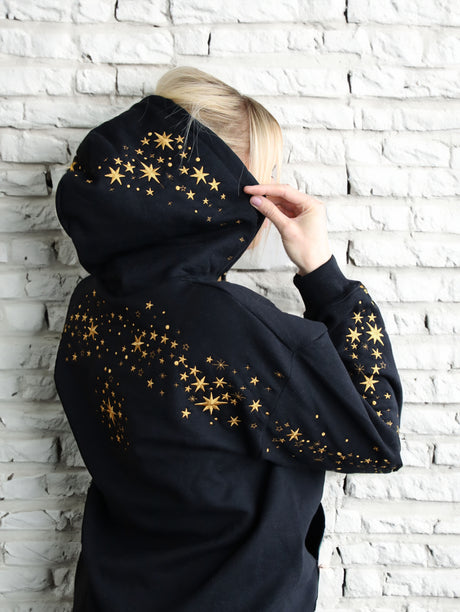 Person wearing a black hoodie with machine embroidered gold star against a white brick wall.