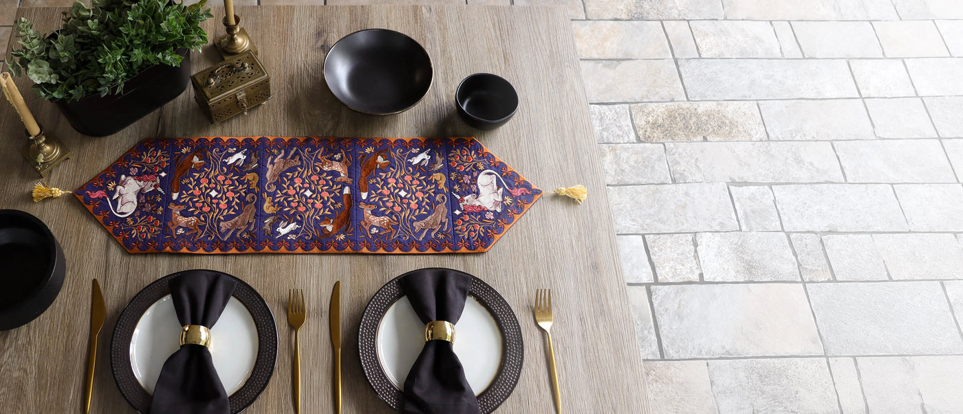 An overhead view of a styled dining table featuring a long embroidered table runner with a blue and gold pattern, black plates with black napkins, gold cutlery, and a light stone tile floor in the background.