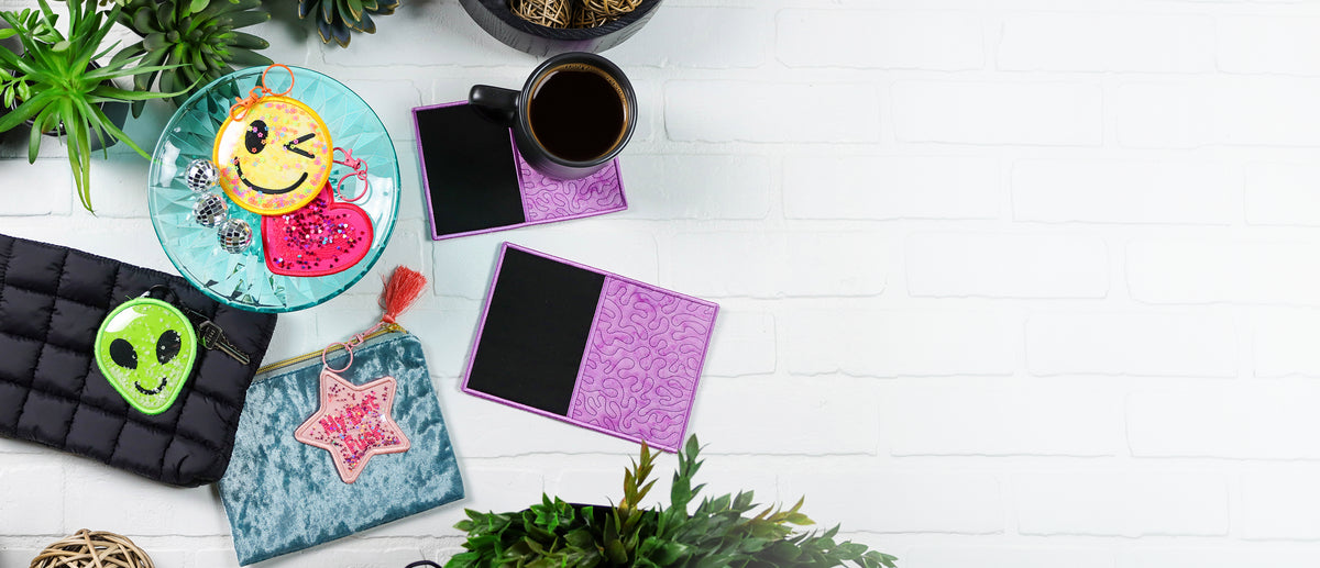 Colorful Y2K glitter shaker keychains and purple in-the-hoop mug rugs displayed with a coffee cup, plants, and accessories on a white tabletop.