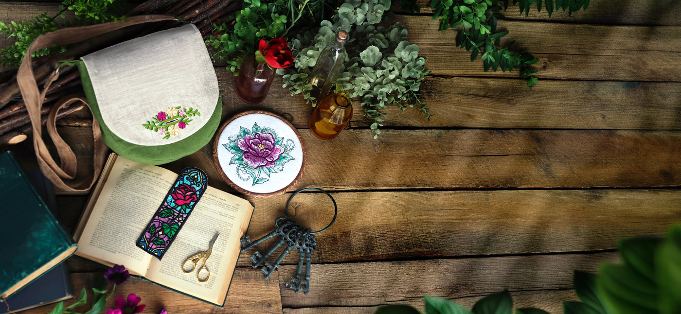Machine embroidery floral buttonhole on purse, rose in hand embroidery hoop and floral bookmark on a wooden surface. 
