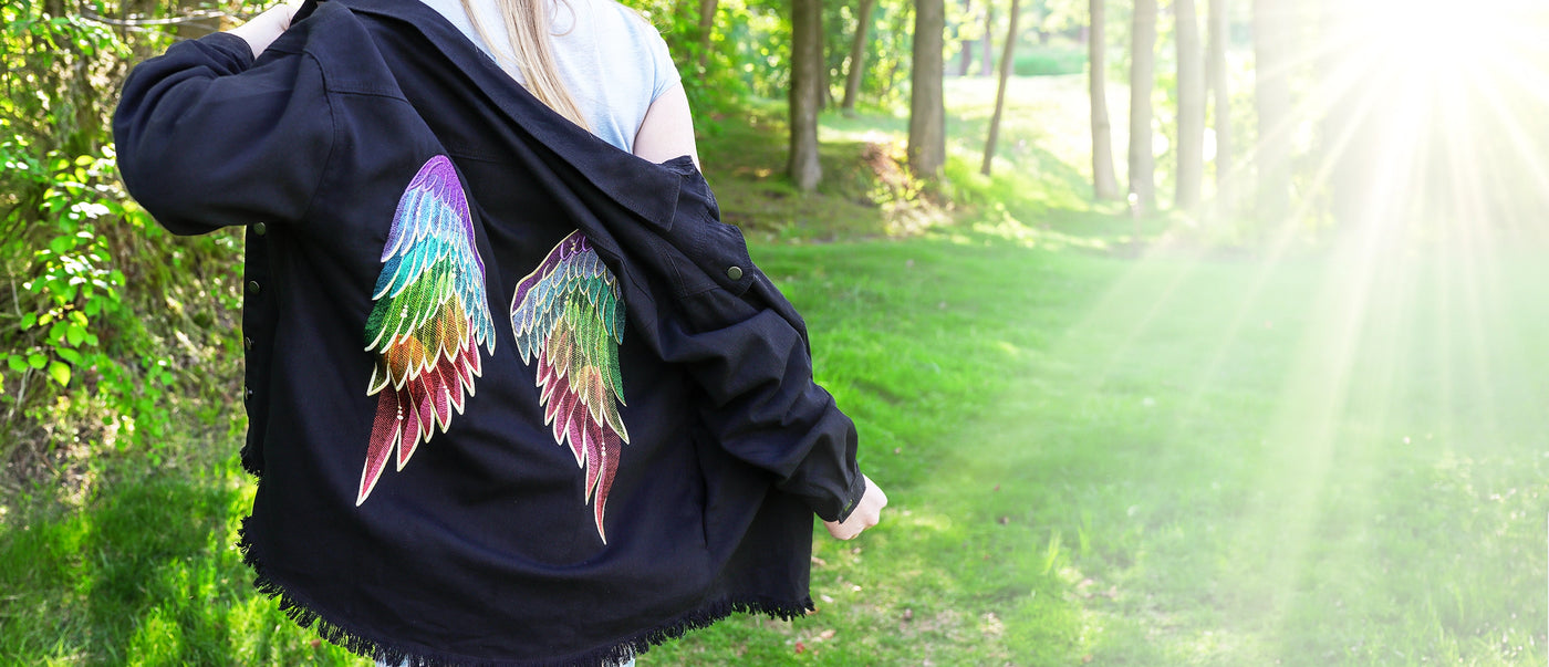 Person wearing a black jacket with machine embroidered rainbow angel wings on a grassy area.