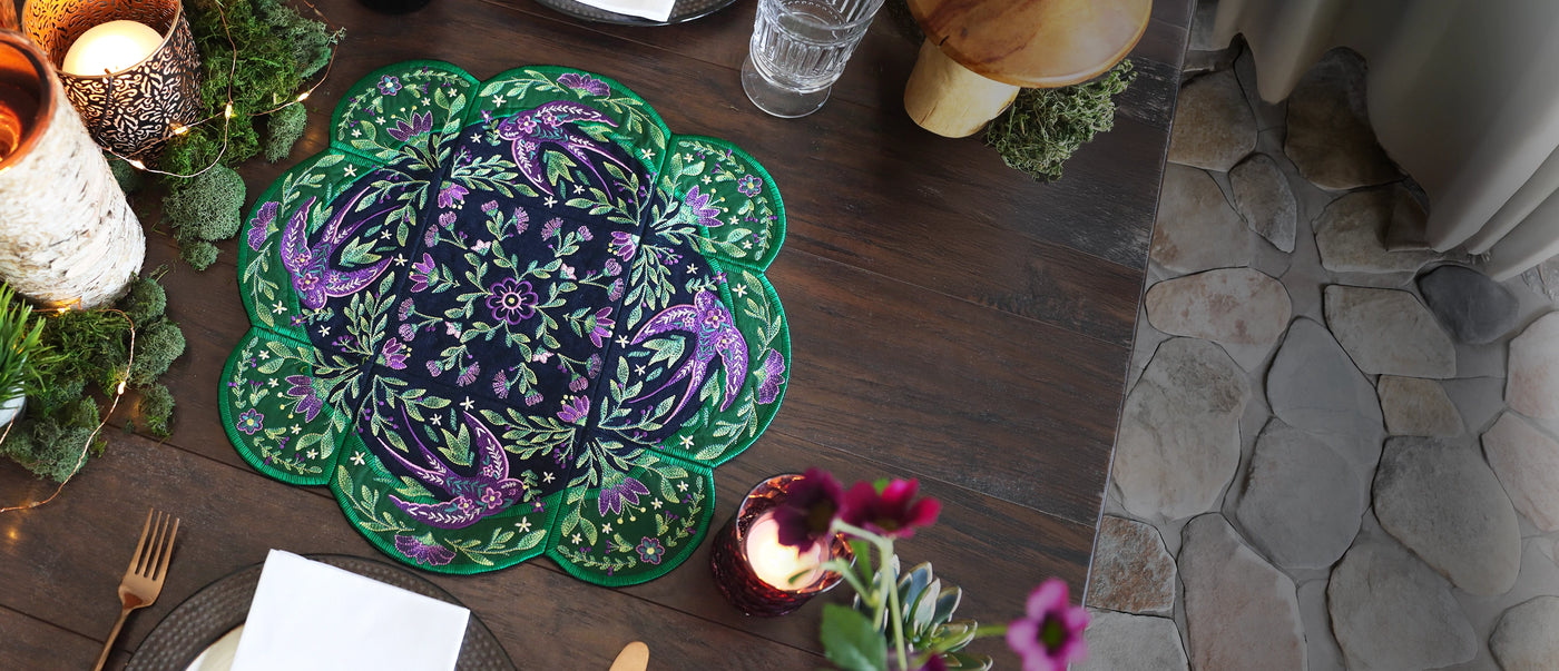 Decorative machine embroidery tabletop design with green and purple flowers and swallows on a wooden table with candles and flowers.