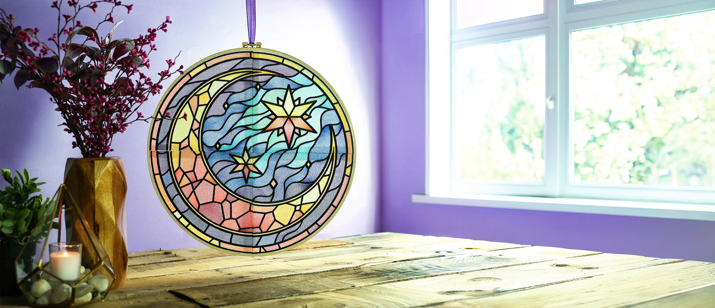 Stained glass style machine embroidered moon and stars suncatcher hanging in a bright window, with pastel colors, flowing sky details, and celestial motifs above a rustic wood table.