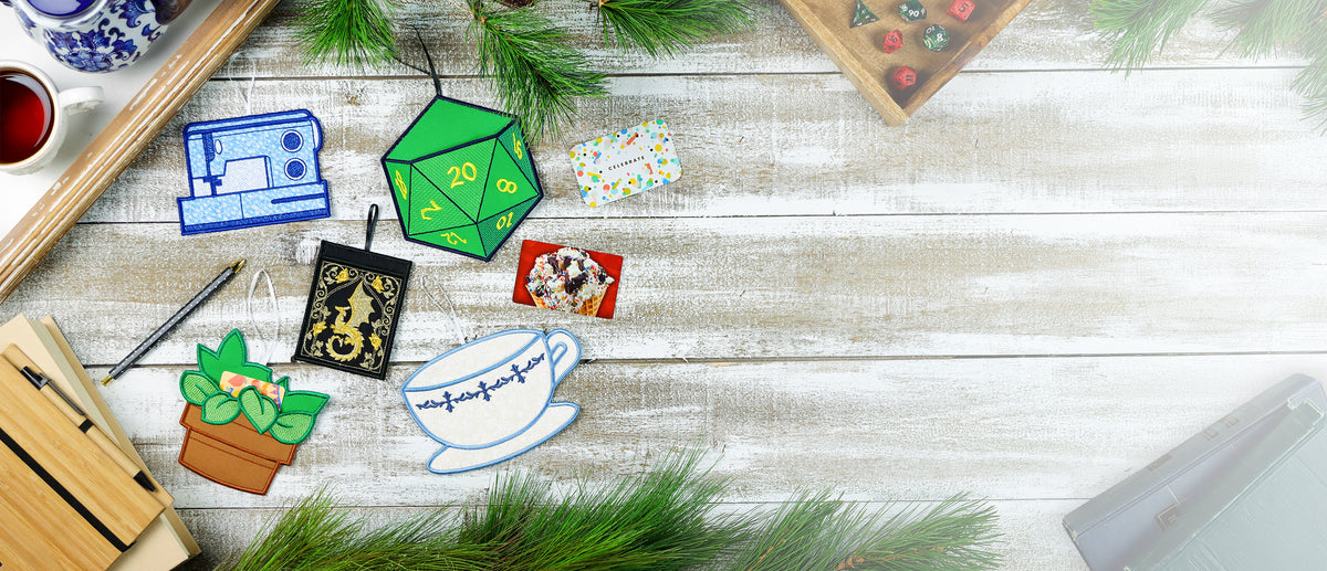 In-the-hoop hobby-themed gift card holders, including a sewing machine, D20 dice, dragon book, potted plant, and teacup, arranged with gift cards on a rustic tabletop.