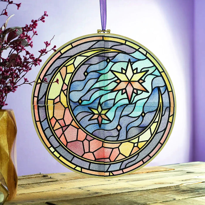Stained glass style machine embroidered moon and stars suncatcher hanging in a bright window, with pastel colors, flowing sky details, and celestial motifs above a rustic wood table.
