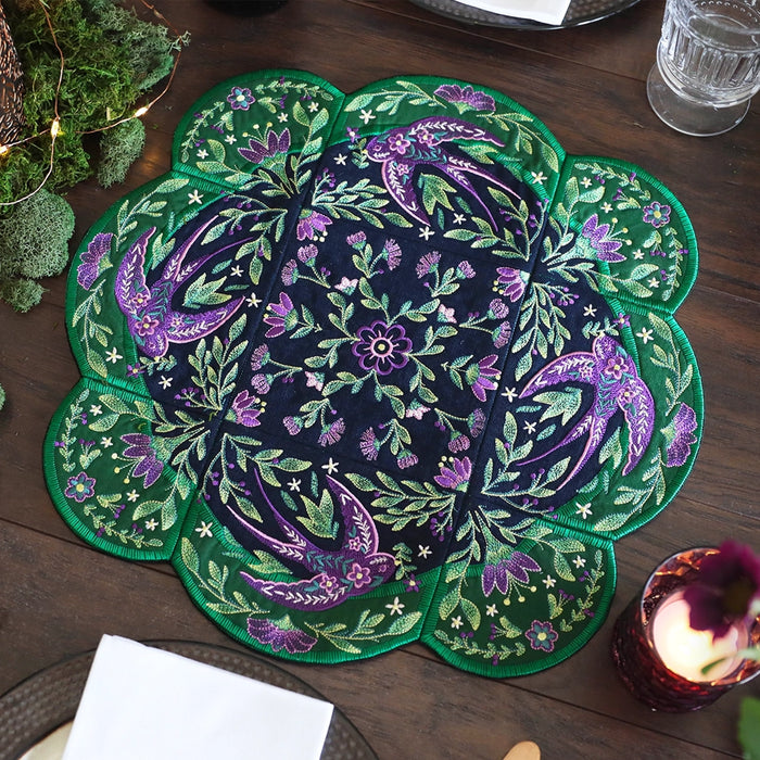 Decorative machine embroidery tabletop design with green and purple flowers and swallows on a wooden table with candles and flowers.