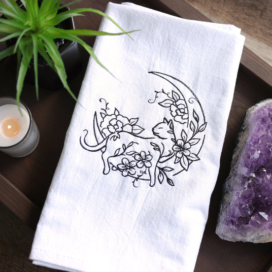 White towel embroidered with a cat & florals design on a wooden tray with a candle and crystal