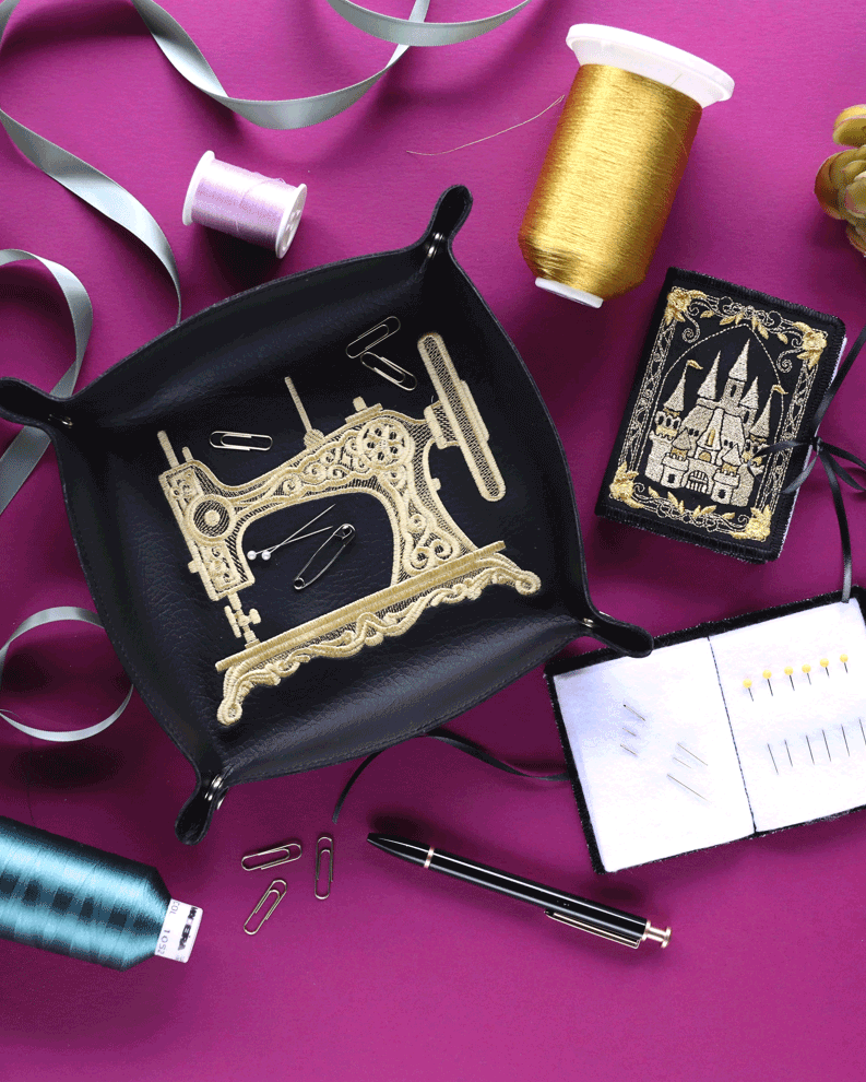 Decorative black leather tray showcasing a vintage sewing machine embroidery and embroidered needlebooks in gold, surrounded by spools of thread, needles, and sewing notions for elegant craft storage or gift ideas