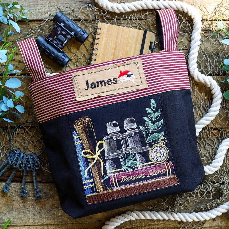 Tote bag with 'James' name tag and 'Treasure Island' bookshelf machine embroidered design on a wooden surface.