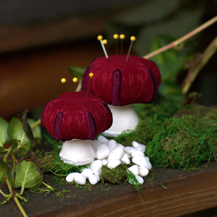 Two red and white machine embroidered pincushion mushrooms on a wooden surface with greenery.