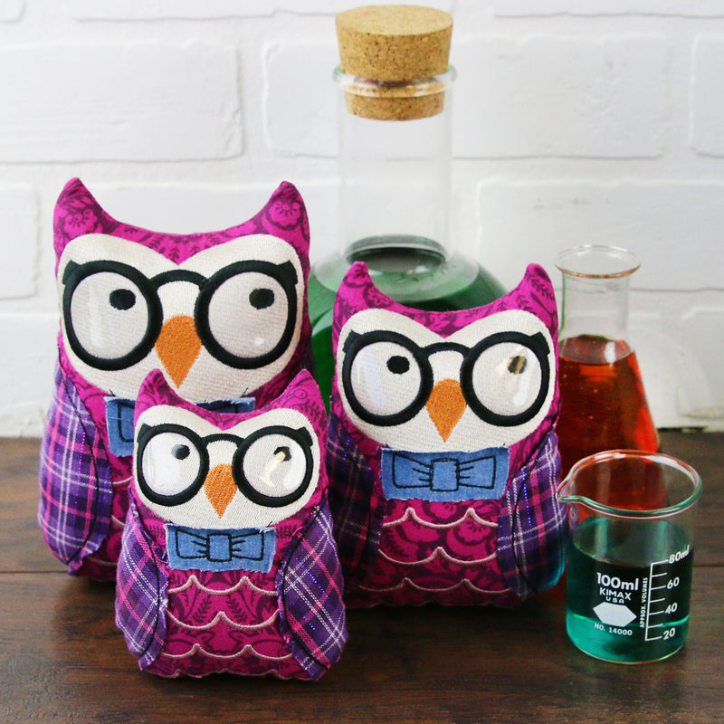 Three machine embroidered in-the-hoop owl stuffies with glasses and bows on a wooden surface.